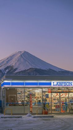 a lawson convenience store stands in the foreground, illuminated against the fading light of dusk. behind it, the iconic snowcapped peak of mount fuji dominates the background, creating a striking contrast between the ordinary and the majestic. the scene evokes a sense of serenity and quiet beauty, capturing the essence of a winter evening in japan.