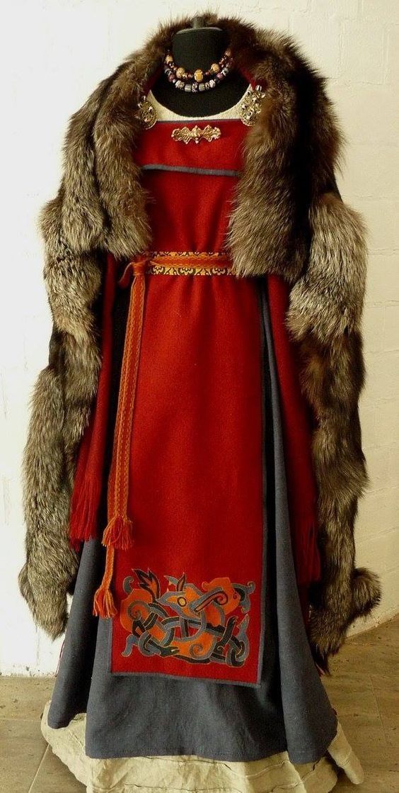 a woman dressed in a viking costume with a red wool dress and a blue skirt, brown fur cloak, and a beaded necklace. the dress is ornamented with embroidery.