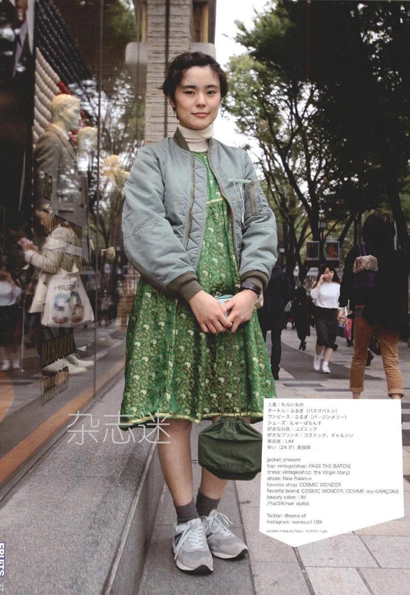 a young woman poses on a city sidewalk in front of a store window. she's wearing a green floral dress, a gray bomber jacket, and gray sneakers. she's carrying a green tote bag and has short, dark hair. the photo has a slightly muted color palette.