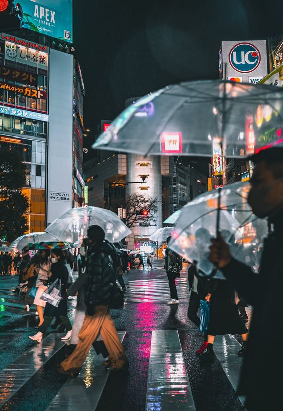 this is a blurry image of a busy city street at night with many people crossing the crosswalk. many people are holding umbrellas, implying it's a rainy night. there are buildings with bright signage on both sides of the street and illuminated signs reflected on the wet asphalt. the perspective is eye level and the lighting is low with neon highlights.  this photo evokes a sense of hustle, excitement and urban energy.