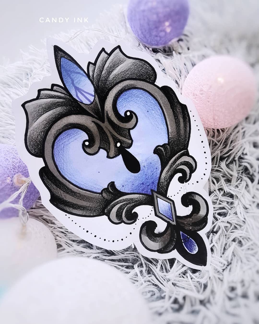 a drawing of a stylized heart shaped tattoo design on white paper with a crumpled texture. the design has a blue crystal at the top and a blue gem at the bottom. it is drawn in a blackwork style with grey shading. the background is white with purple and pink balls out of focus.