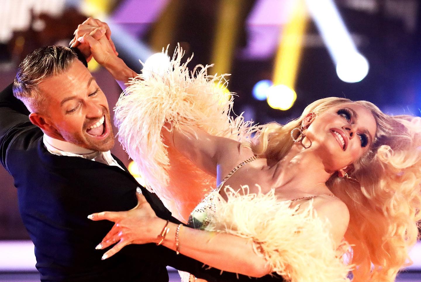 two people are dancing on a brightly lit stage. the person on the left is a man with short blonde hair, wearing a black suit and a white shirt. he is smiling broadly.  the person on the right is a drag queen with elaborate blonde hair and makeup, wearing a glamorous costume with feathers and sequins. she is also smiling. they appear to be performing a ballroom dance.