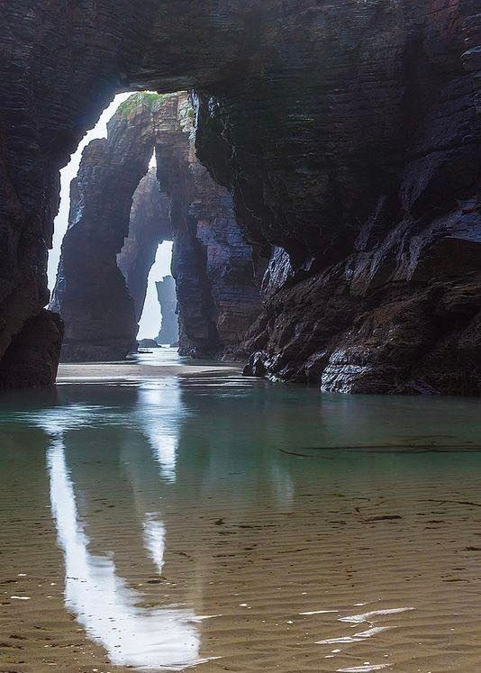 the image captures a scene at playa de las catedrales in ribadeo, lugo, galicia, spain. it features natural rock formations resembling cathedral arches, reflecting in the shallow water on the sandy beach. cliffs surround the area, leading to the sea or ocean. the tide is low, revealing the coastline and allowing access to these unique natural arches. the scene emphasizes the beauty of nature and its attraction for tourism and travel.
