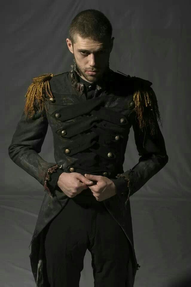 a man stands in a dimly lit setting, wearing a black militarystyle coat with gold epaulettes and multiple rows of buttons. he has short hair and a beard, and his hands are clasped in front of him. the coat appears worn or distressed, adding to a dramatic or theatrical effect.