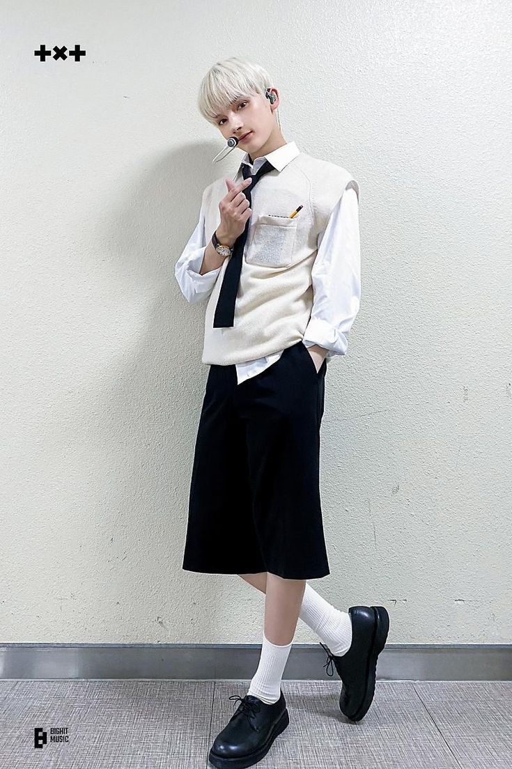 a fulllength shot of a young man in a formal outfit. he is wearing a white longsleeved shirt, a creamcolored sweater vest, a black tie, black culottes, white socks, and black dress shoes. he has short, light blonde hair and is making a small heart gesture with his fingers. he also has a microphone attached near his mouth. the background is a plain white wall and a gray floor. the logo "txt" is in the top left corner, and the "big hit music" logo is in the bottom left corner.