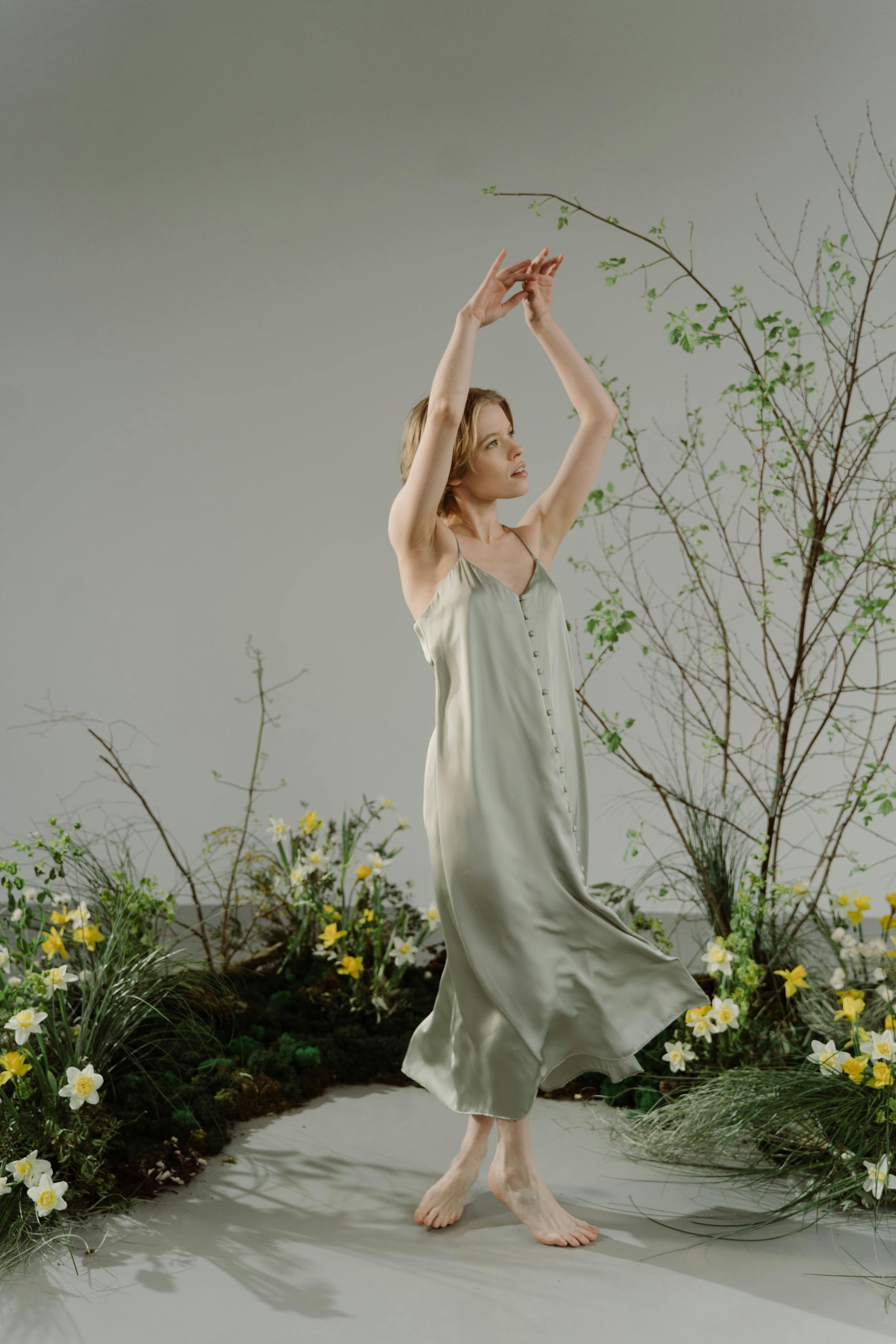 a young woman in a flowing, sage green silk slip dress stands barefoot amidst a spring floral arrangement. her arms are raised gracefully, and her pose conveys a sense of serenity and peace. the backdrop is a minimalist light grey studio setting, enhancing the ethereal quality of the image. the overall aesthetic is delicate and gentle, with soft lighting illuminating the scene. the flowers add a natural touch, complementing the dress's soft color and creating a harmonious atmosphere.