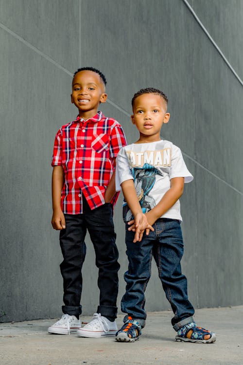 two young african american brothers stand sidebyside against a gray wall.  the boy on the left wears a red plaid shirt and dark jeans, while the boy on the right wears a white batman tshirt and blue jeans.  both boys are smiling and appear happy.  they are wearing sneakers and sandals, suggesting a casual, everyday setting.  the overall feel of the image is warm, friendly, and captures a moment of sibling camaraderie.