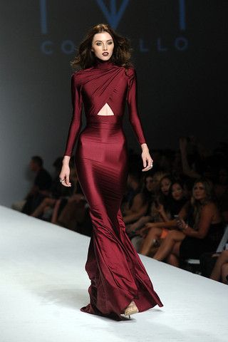 the image shows a slender female fashion model walking down a runway during a fashion show. she's wearing a stunning, long, formfitting, dark red (burgundy) gown with a high neckline and a subtle cutout detail at the waist.  the dress has a mermaid or fishtail style skirt. the model's hair is styled in loose waves, and her makeup is fairly neutral. the background is blurred, showing other individuals seated in the audience watching the fashion show.  the background also displays the name of the fashion house or designer. the overall feel is one of elegance and high fashion.