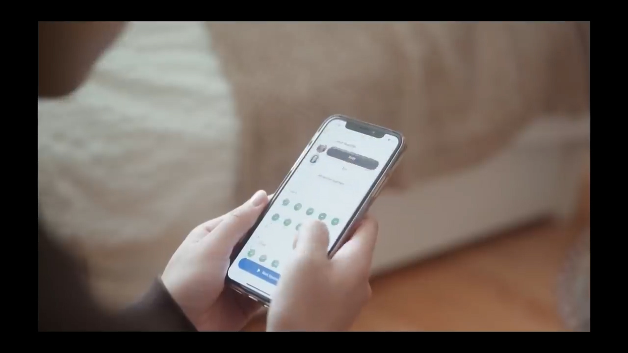 a closeup shot of a person holding a smartphone in their hand. the screen is lit up, displaying a social media app with profile pictures and buttons. the background is blurry, suggesting a bedroom setting. the lighting is soft and natural.