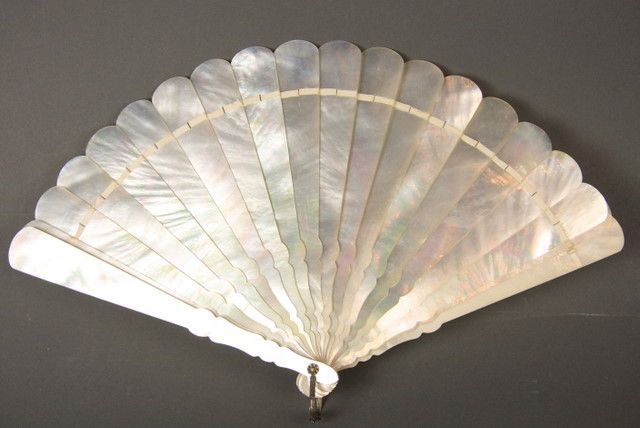 an ornate hand fan made of iridescent motherofpearl segments is displayed against a dark background. the fan is open, revealing its scalloped edges and the shimmering, pearlescent quality of the material. a metal fastening secures the base of the fan.
