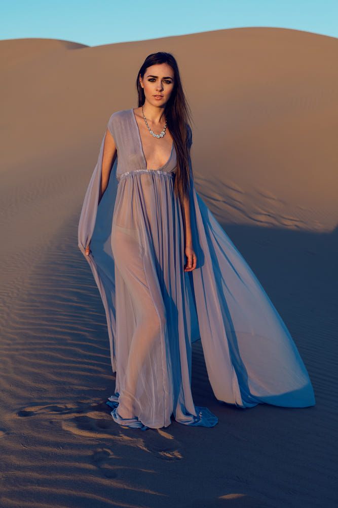 a woman in a flowing, sheer, periwinkle blue dress stands on a sand dune. the dress has a deep vneckline and flowing sleeves. she is wearing a delicate necklace. the sunlight is soft and casts a warm glow on the sand.  the background is a serene landscape of rolling sand dunes.