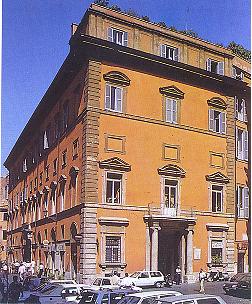 the image shows a tall, orange building in an urban setting, likely in europe, potentially italy or rome. the building features classical architecture with windows, columns, and decorative elements on its facade. there are cars parked in front of the building and people walking on the street. the building appears to be historic and wellmaintained.
