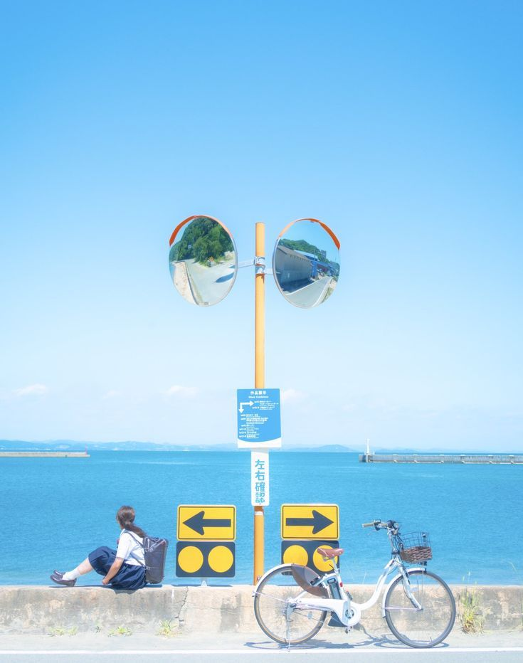 a girl sits on a low concrete wall overlooking the ocean on a bright sunny day. next to her is a bicycle parked near a traffic mirror and road signs. in the background is a view of the ocean, a distant island, and a pier, all under a clear blue sky.