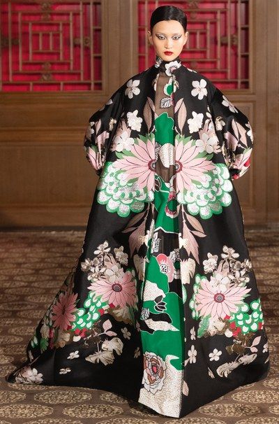 a full shot of a female model showcasing a floorlength gown. the dress is predominantly black with a striking floral pattern featuring pink, green, and metallic accents. the design appears to be inspired by asian aesthetics, with intricate details and a luxurious, almost brocadelike texture. the model's makeup complements the dress' colors, and she stands against a backdrop suggesting a fashion show or photoshoot setting. the overall impression is one of high fashion and elegance.