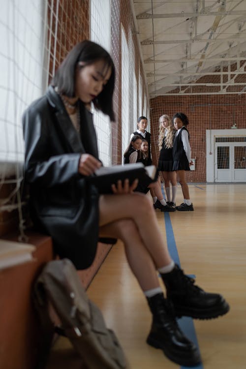 a young woman sits on a bench in a school hallway reading a book. a group of schoolgirls in uniform stand behind her in the background. the overall mood is casual and friendly.