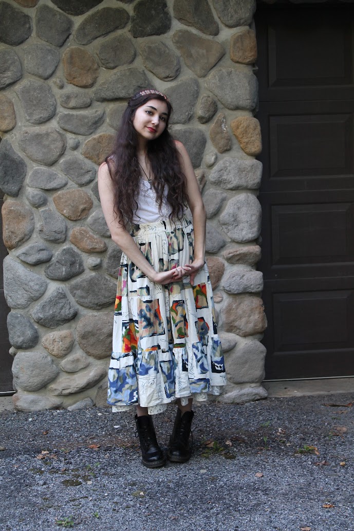 a young woman with long brown hair, wearing a headband, stands against a stone wall. she's dressed in a long, flowing maxi skirt with a patchwork design, a white tank top, and black doc martens boots.  the background includes a dark brown garage door.  she is posing for a portrait, looking directly at the camera. the overall style evokes a bohemian or vintage aesthetic.