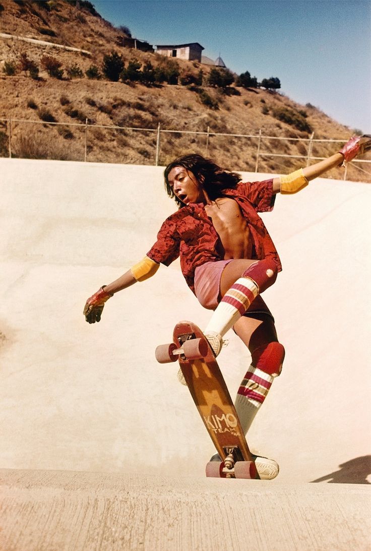 a young man in 70s skateboarding attire is in midair while performing a trick on his skateboard. he is wearing knee pads and gloves. he is flying over a ramp in a halfpipe. the ground is concrete and there are hills and grass in the background. it's a sunny day, with a blue sky.