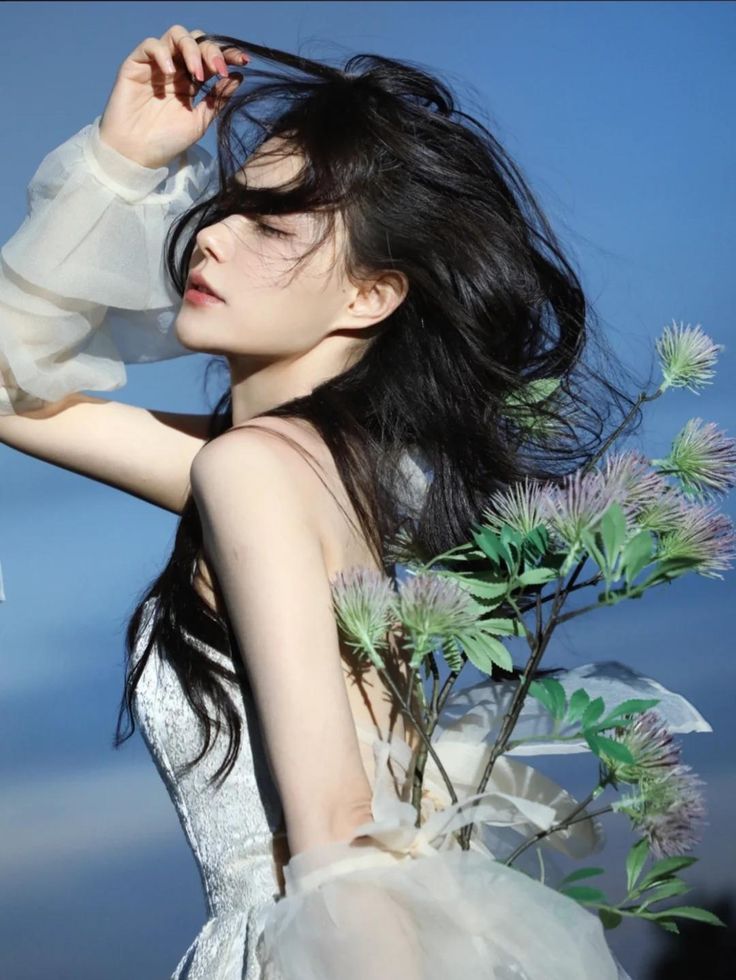a beautiful woman with long black hair is posing outdoors in a white dress with delicate flowers in her hand. the blue sky is in the background. the woman is looking away from the camera, her expression is soft and dreamy.