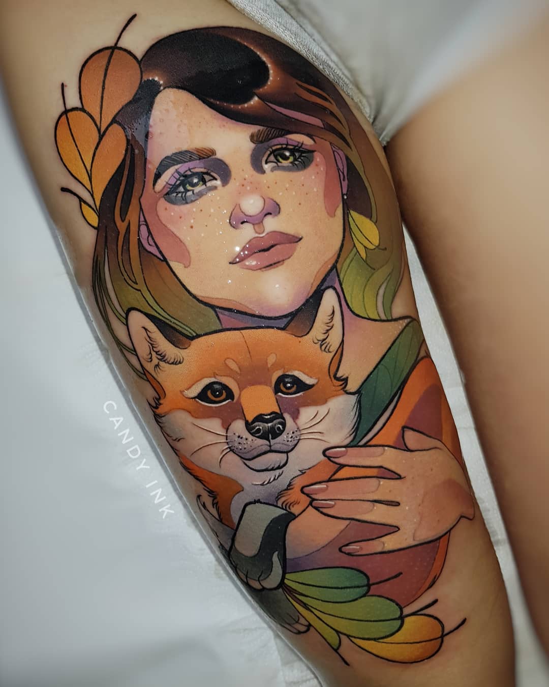 a neotraditional tattoo on someone's upper thigh. it features a portrait of a woman with her pet shiba inu nestled in her arms. the woman and the dog are framed by colorful abstract shapes resembling leaves or feathers. the colors in the tattoo are mostly warm and earthy tones like browns, oranges, and yellows. the artist's signature "candy ink" is visible on the left side of the image.