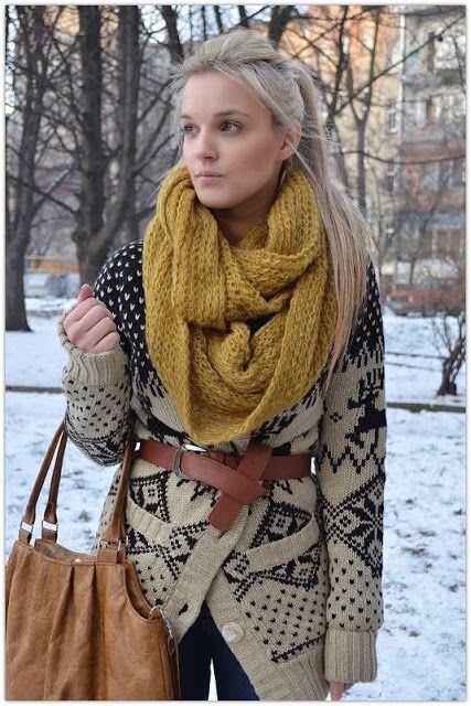 a blonde woman is posing for a portrait outdoors in the winter. she is wearing a yellow scarf, a black and white patterned cardigan, a brown belt, a brown bag, and blue jeans. the background is blurry with trees and snow.