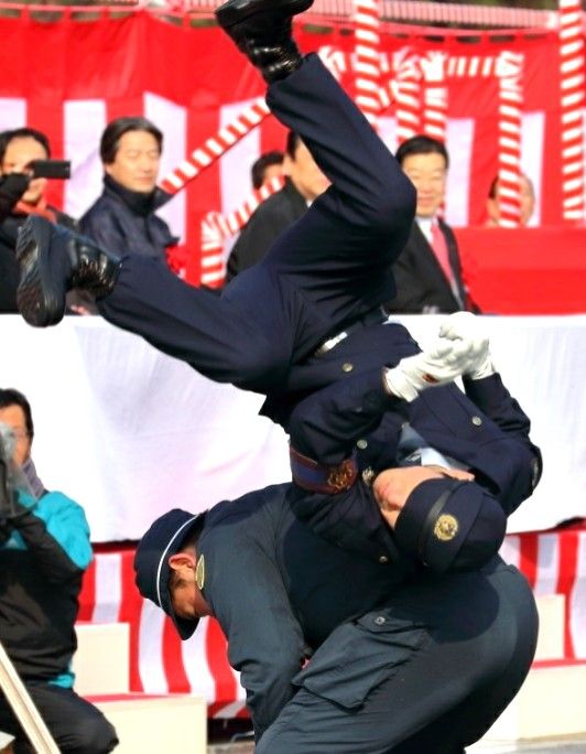 a police officer is shown tackling a suspect in a street, during a public event. the officer is wearing a blue uniform and is in midair. the suspect is wearing black pants and a blue jacket. both are in the middle of a takedown, with the police officer's hand near the suspect's neck.  they appear to be surrounded by other people and a red and white striped banner.