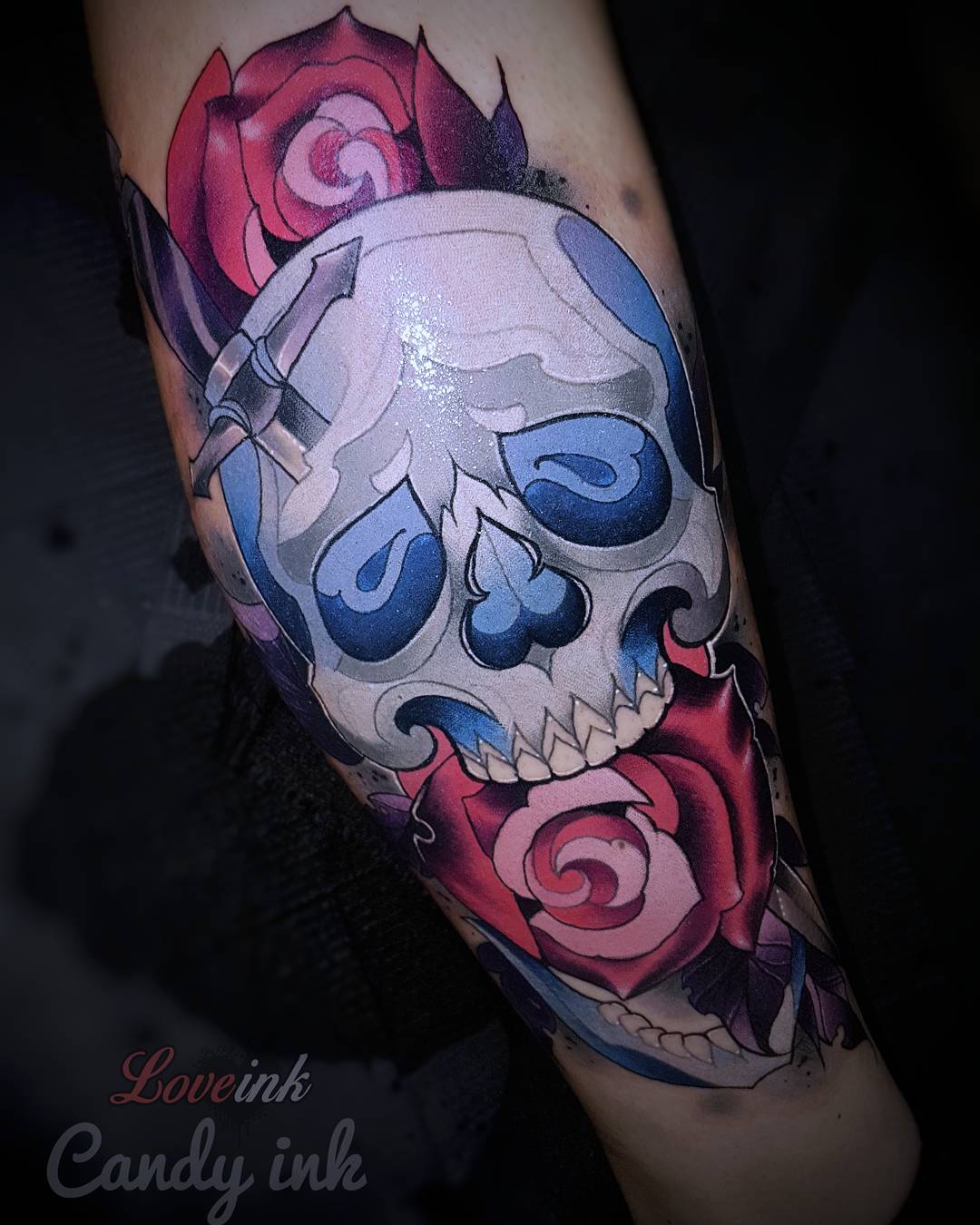 a tattoo of a skull with a dagger through it and roses around it. the skull is white with blue details, and the roses are red and pink. the tattoo is on someone's arm.