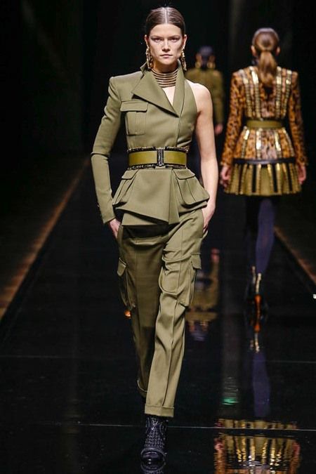 the image showcases a female model on a runway, exhibiting a stylish militaryinspired outfit. the ensemble features an olivegreen oneshoulder top with a peplum waist, paired with matching cargo pants. a gold belt accents the waist. the model also wears highneck choker and statement earrings, complementing the outfit's sophisticated edge.  her footwear consists of dark ankle boots. the background is a dark runway, hinting at a fashion show. another model in a contrasting gold and animal print outfit is visible in the background, blurred, indicating a fashion show setting. the overall aesthetic is sharp, elegant, and ontrend, combining military influences with sophisticated detailing.