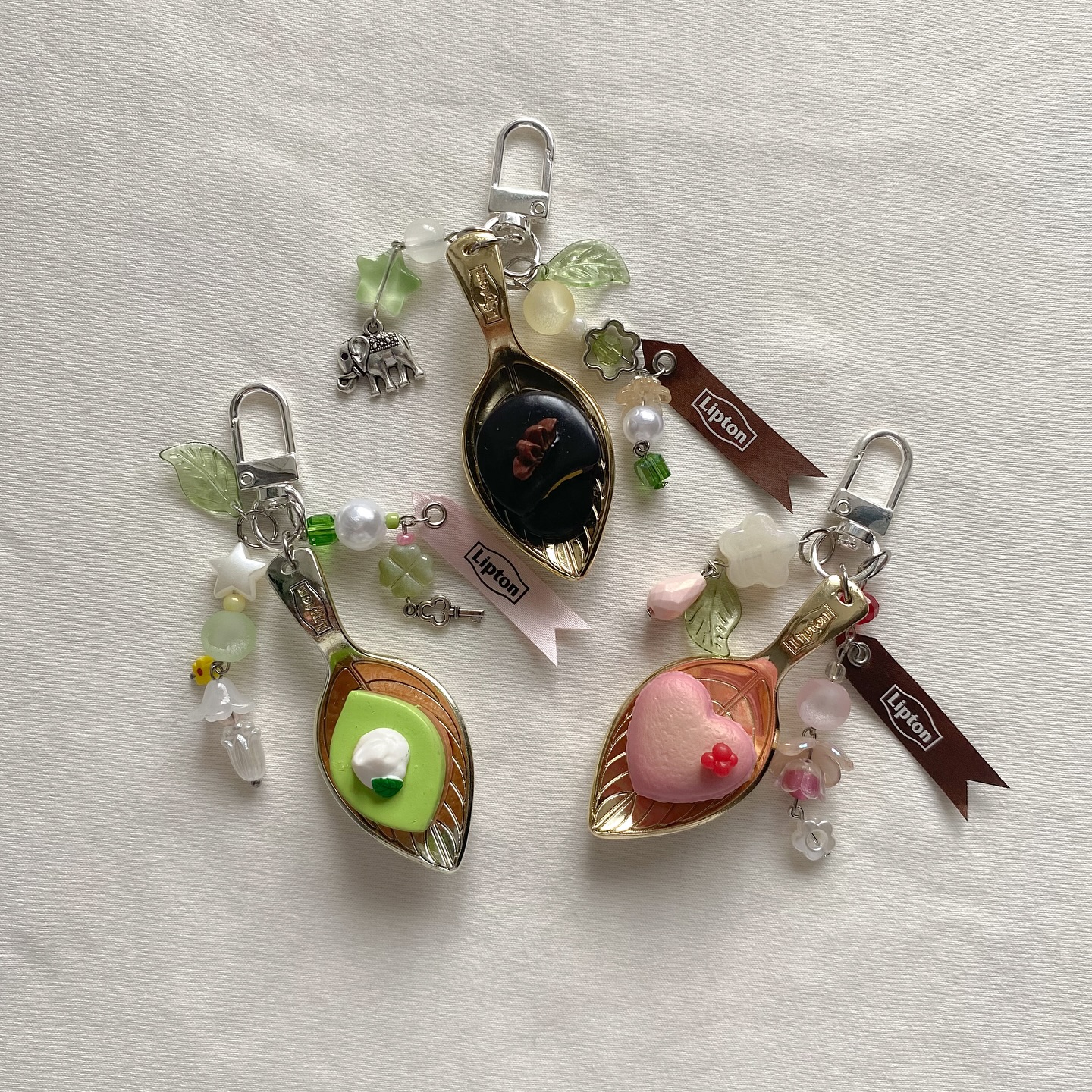 the image showcases a collection of three handmade keychains featuring repurposed spoons as a base, each adorned with various kawaiistyle decorations. one keychain features a kiwi fruit slice made of polymer clay in the spoon, another has a chocolate disc, and the third has a pink heart. all three keychains have lipton brand ribbon tags, various beads, flowers, and leaf charms attached to the keychain loop. the keychains are photographed on a neutral fabric background.