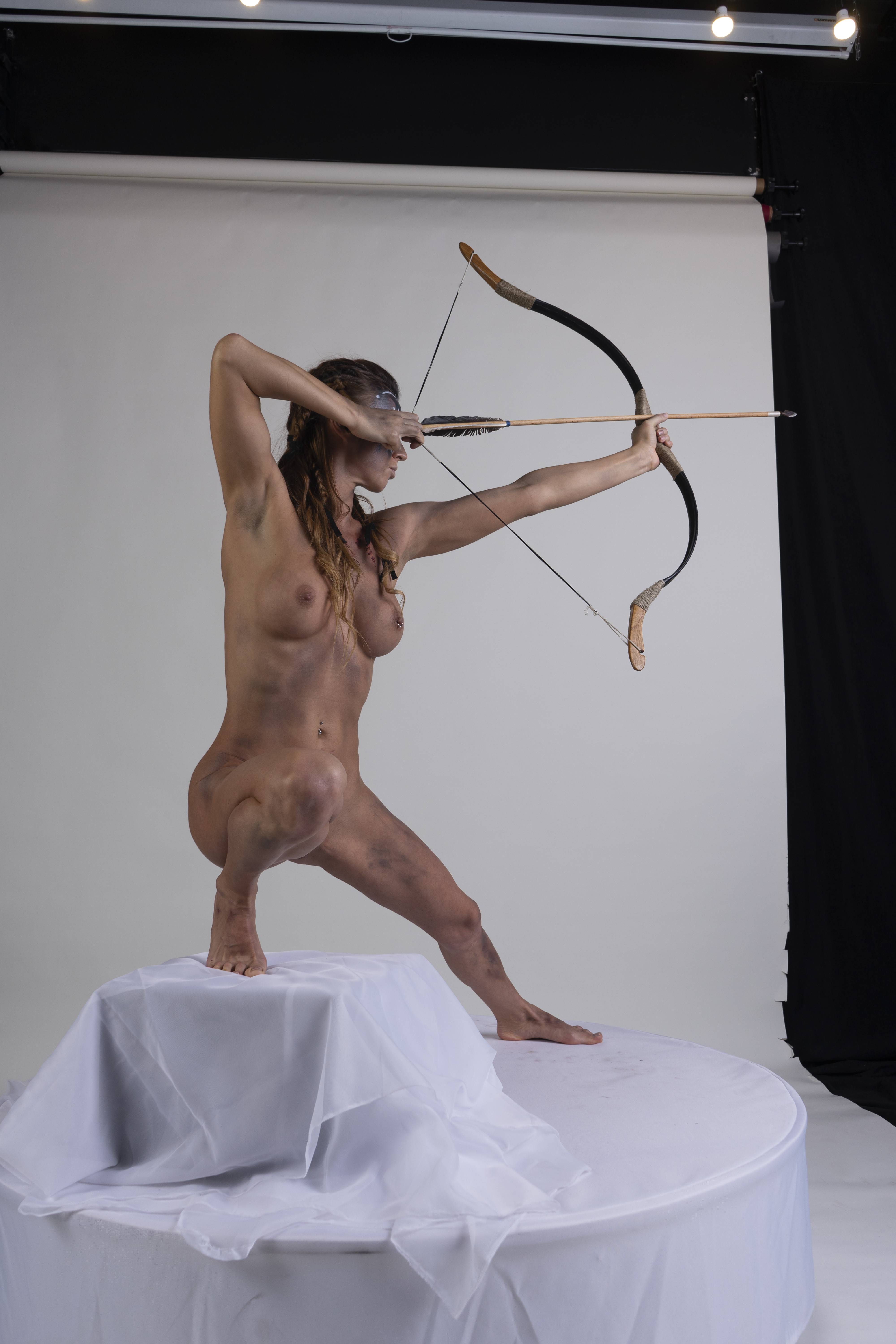 a nude woman with tribal makeup on her face is posed as an archer in a studio setting. she is kneeling on a white platform covered with white fabric. she is holding a bow and arrow, aiming forward. the background is a plain white backdrop with a black curtain visible on the right.