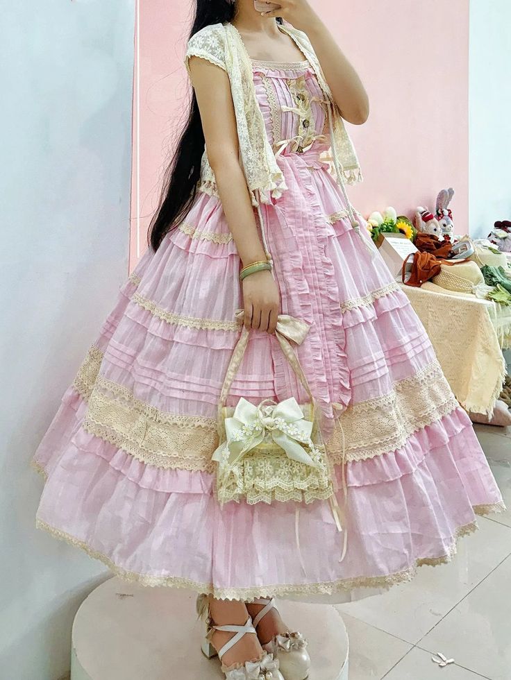 a person wearing a pink lolita fashion dress is standing indoors. the dress is long and full with multiple layers of pink fabric and lace trim.  a matching cropped cardigan with lace trim is worn over the dress.  the person is also carrying a cream colored purse with a bow on it. the photo is taken from the chest down,  and only the person's torso and lower body are visible.