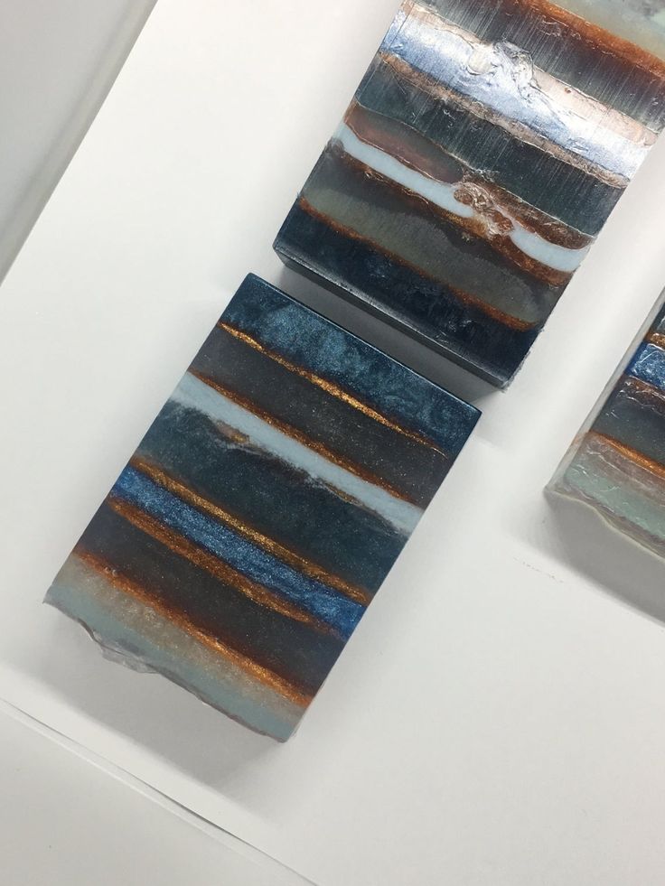 two pieces of handmade soap with abstract blue, green, brown, white, and gold stripes are shown. the soaps are laid out on a white background and have a natural and luxurious feel. the image is welllit and in focus, showcasing the intricate details of the soap's design.  this type of soap would be a perfect gift for someone who enjoys a luxurious spa experience.