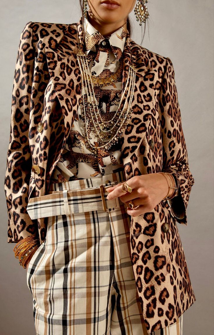 loseup view of a fashion model wearing a chic outfit. the model is adorned in a leopardprint blazer, paired with beige and brown plaid trousers. she's accessorized with a multitude of gold necklaces, large statement earrings, bangles on her wrists, and several rings. the overall style is sophisticated and luxurious. the background is a simple, muted beige tone which emphasizes the model's outfit.  the image is welllit and professionally styled for a highfashion lookbook or editorial.