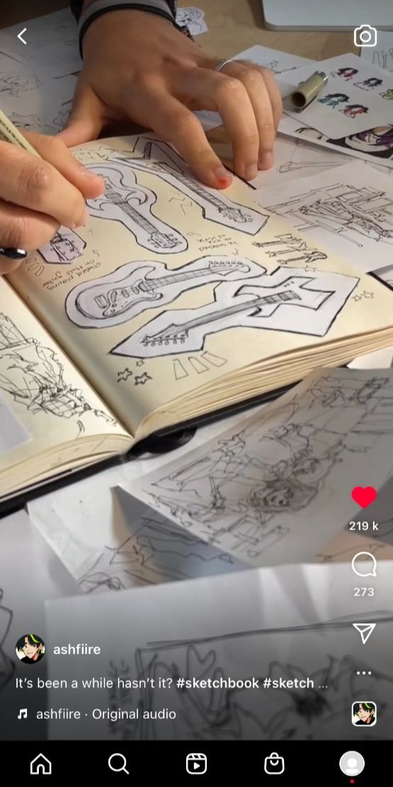 a person is drawing a guitar in a sketchbook. they are holding the sketchbook open with their left hand and drawing with their right hand. the sketchbook is on a table and there is another piece of paper on the table next to it. the person is wearing a black shirt.