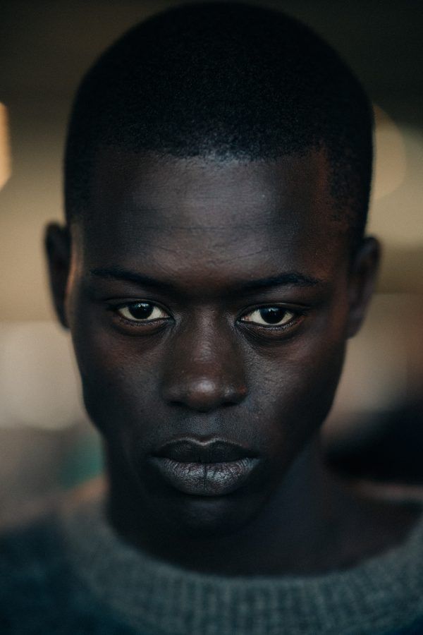 a closeup portrait of a young, darkskinned man with a serious expression. he is wearing a sweater and looking directly at the camera with an intense and confident gaze. the image highlights his striking features and beautiful skin.