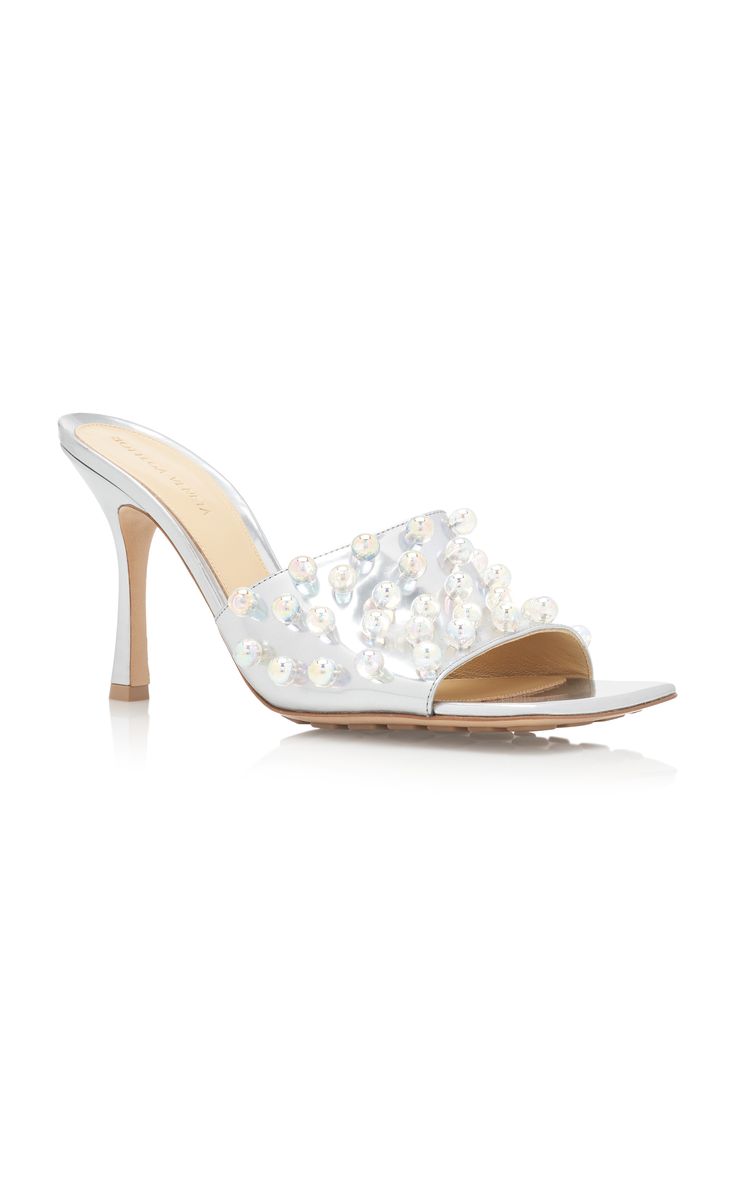 a pair of silver amina muaddi mules are shown on a white background. the shoes are high heels with a square toe and open back. they are embellished with iridescent pearls across the top of the foot.  the shoes are angled slightly to the right.  a subtle reflection of the shoes is visible below.