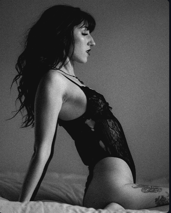 the black and white photograph shows a woman with long dark hair, sitting on a bed in a black lace bodysuit. her profile is visible, and her gaze is directed downward. the image has a sensual and intimate mood, enhanced by the use of shadows and the monochromatic palette. the woman is adorned with a tattoo on her leg, and the overall aesthetic is elegant and artistic. the image is suggestive of fashion or beauty photography.