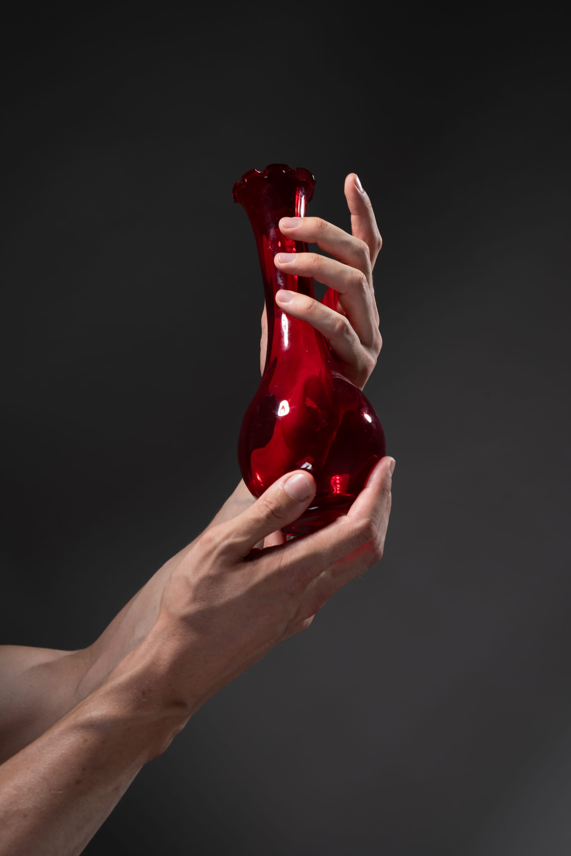 a closeup image of a pair of male hands holding a red glass vase in a studio setting. the vase is transparent, allowing light to pass through it and highlighting its vibrant color. the background is a dark gray, drawing attention to the vase and hands.