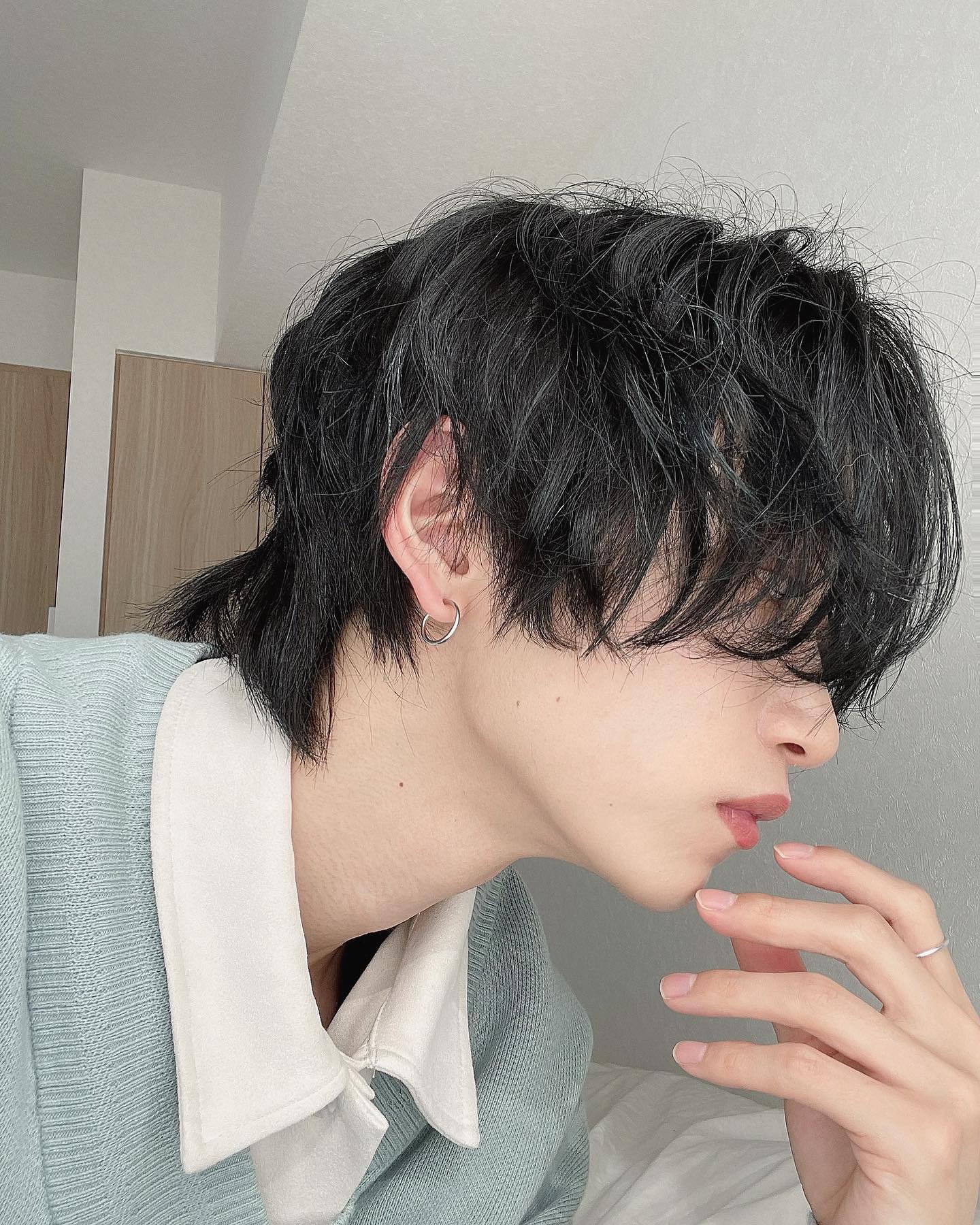 a young man with a trendy, textured mullet hairstyle poses for a closeup selfie. he is wearing a light teal knit sweater layered over a white collared shirt. his head is turned to the side, offering a profile view, and he gently touches his chin with his hand. the photo is taken indoors with soft, natural lighting, creating a serene and stylish atmosphere.