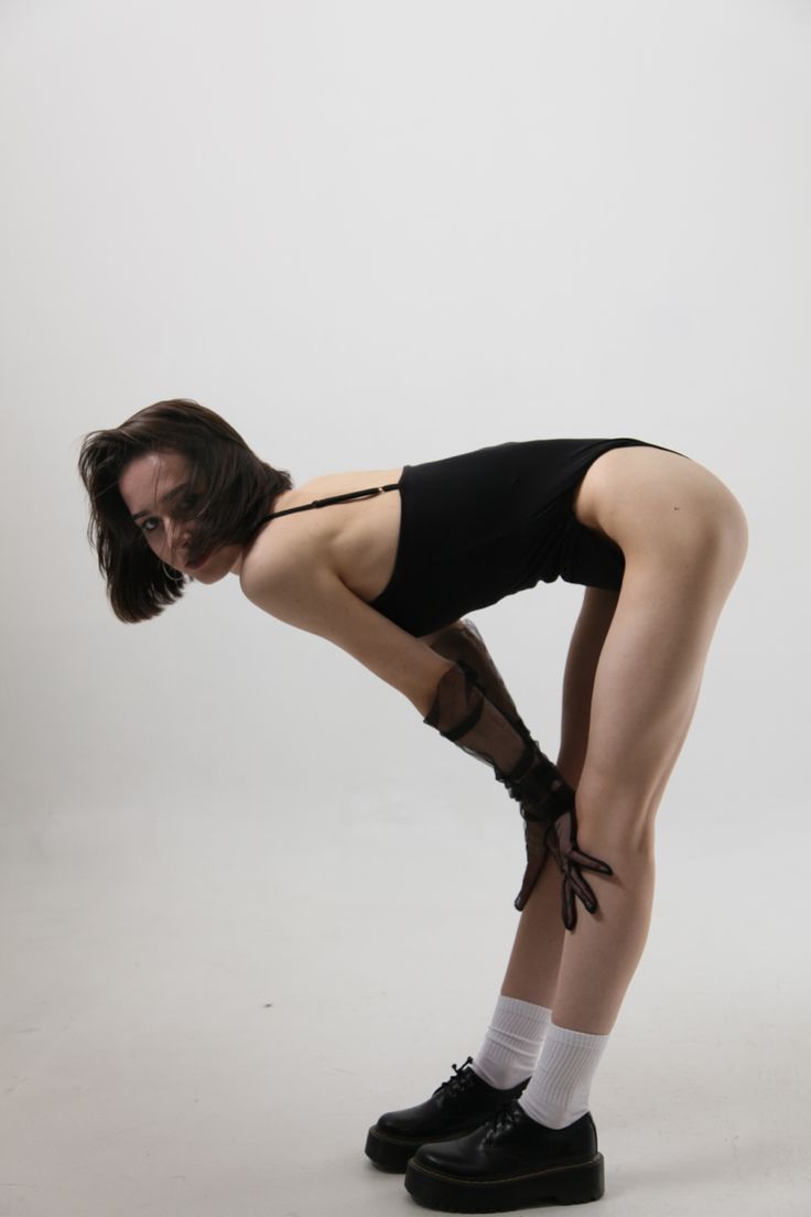 a woman with short brown hair is leaning forward in a studio shot. she is wearing a black bodysuit, sheer black gloves, white socks, and black platform shoes. her hands are resting on her legs. the background is a plain white color.