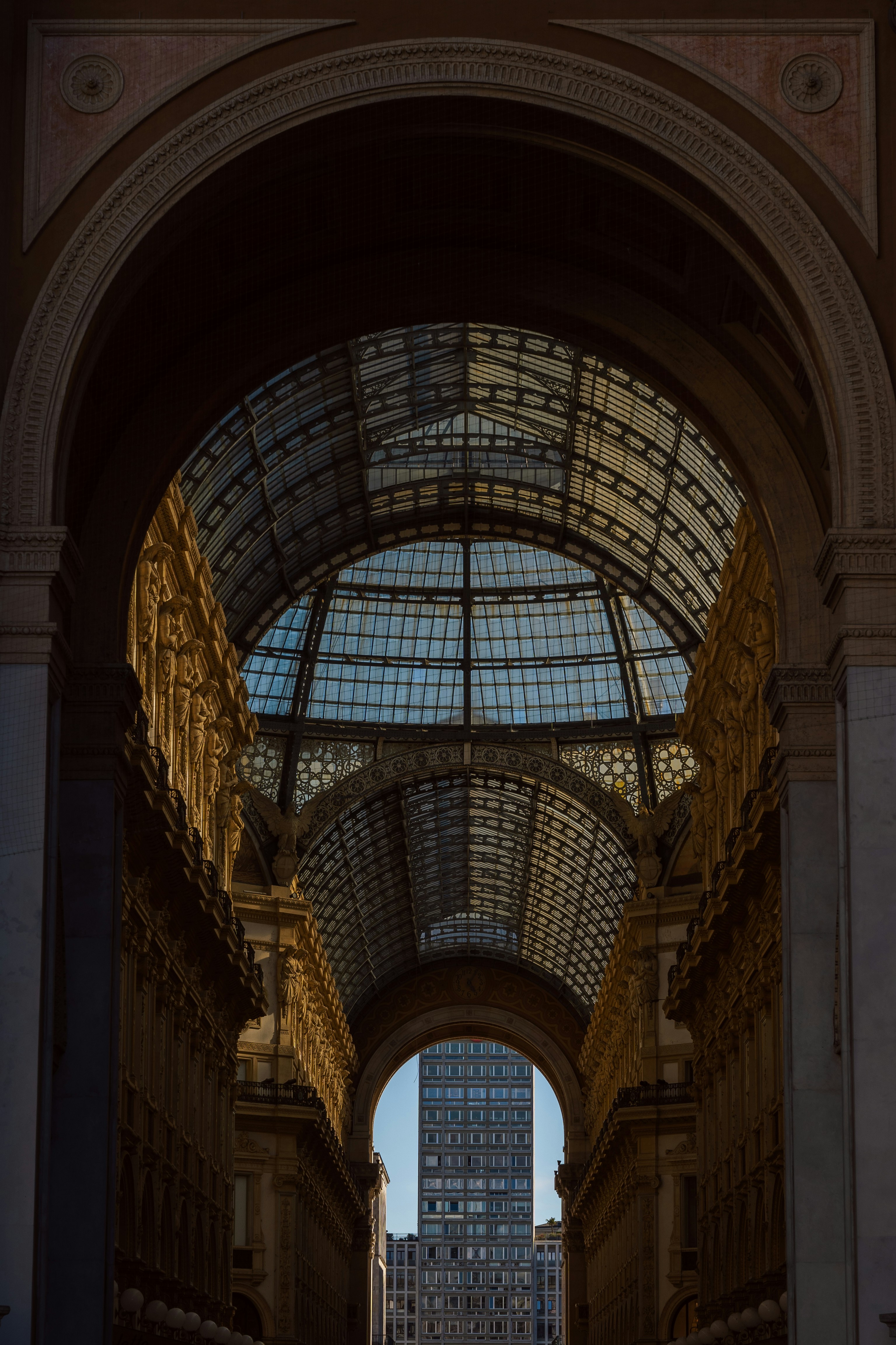 the image captures the grandeur of the galleria vittorio emanuele ii in milan, italy. the perspective is from within the arcade, looking towards one of its iconic glass domes. the sunlight streams through the intricate metal and glasswork of the dome, illuminating the ornate details of the surrounding architecture. the warm, earthy tones of the stone and stucco create a sense of history and elegance. through the archway, a glimpse of a modern highrise building can be seen, showcasing the contrast between the old and new in this vibrant city.