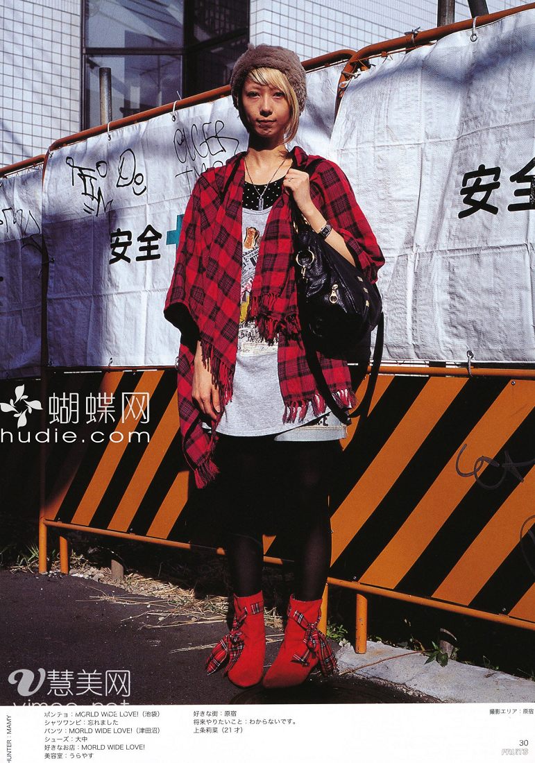 a young woman is posing in front of a fence with japanese writing. she is wearing a red and black plaid shawl, a white tshirt, a black skirt, and red and black plaid boots. she has short, bleached blonde hair and is carrying a black purse. the photo is captioned with japanese text.