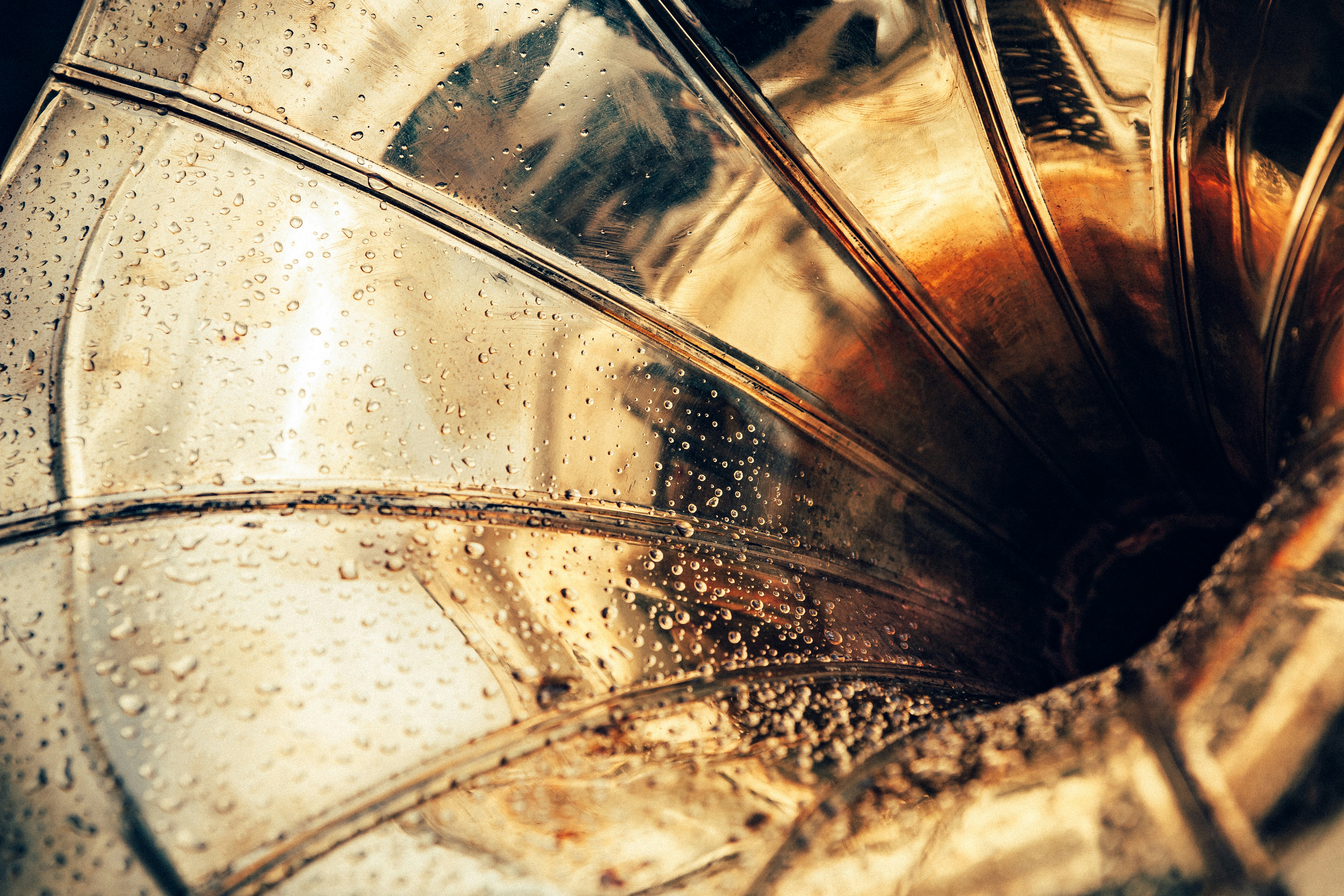 a closeup image of an old gramophone horn with water droplets on its surface. the horn is made of brass and has a shiny, reflective surface. the ridges of the horn are clearly visible, creating a pattern of light and shadow. the water droplets add a sense of texture and depth to the image. the overall tone of the image is warm and nostalgic, evoking a sense of the past.
