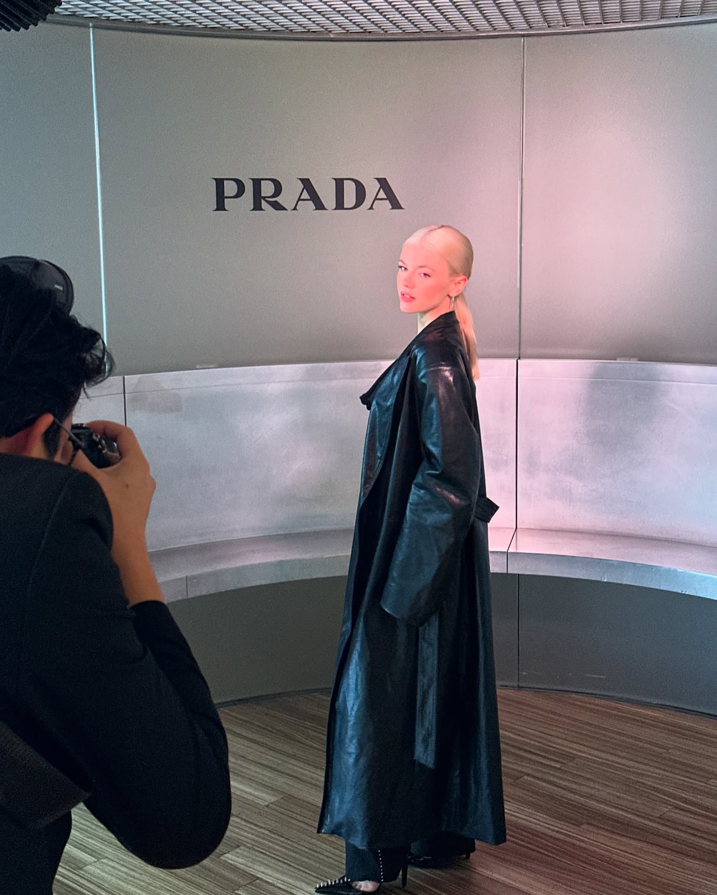 a model poses for a photographer in front of a wall with the prada logo. the model is wearing a long black leather coat and heels. the photographer is taking a picture of the model with a camera.