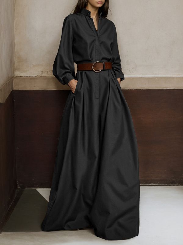 a woman is standing in a long black dress with a long sleeve and a belt. she is posing for the camera and the background has a wall and floor.  the dress is elegant and sophisticated.