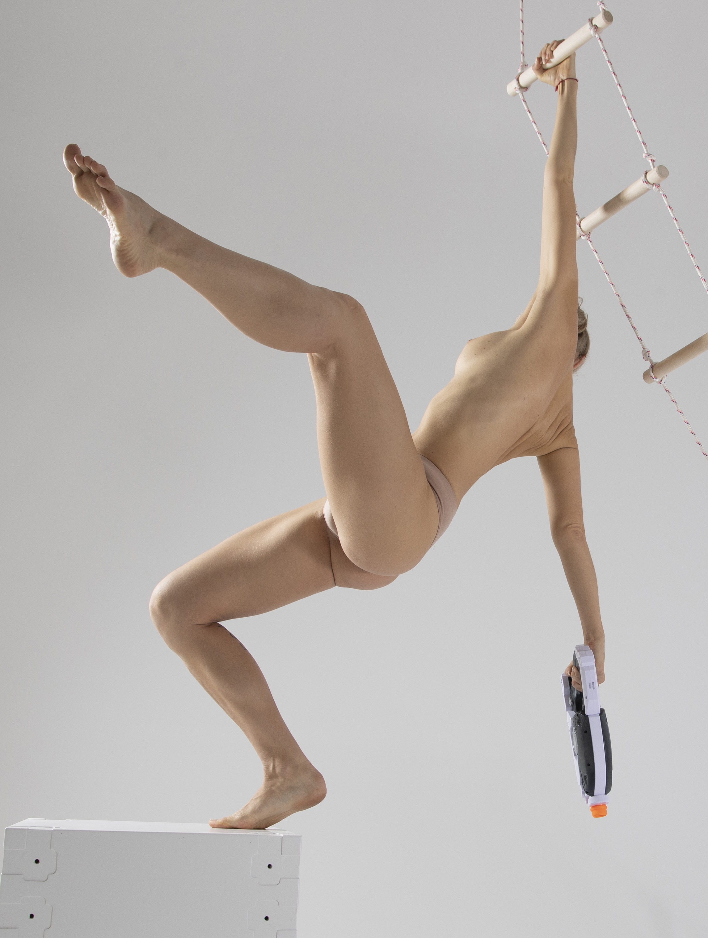a nude woman is hanging from a ladder with one arm, while holding a water gun in her other hand. one foot is resting on a white box and the other leg is extended in the air.