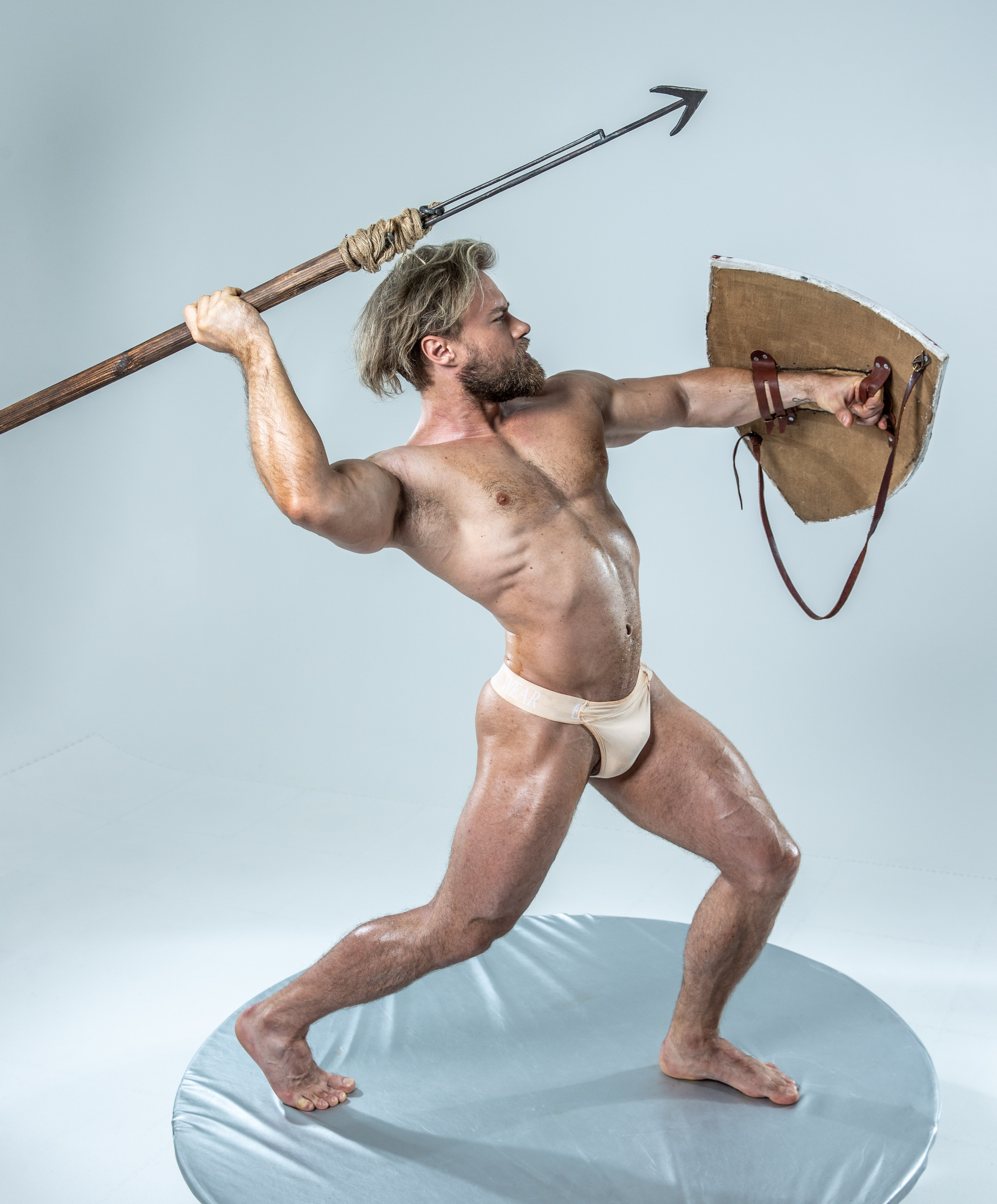 a muscular man, stripped to his underwear, adopts a warrior's pose against a plain grey studio backdrop. he brandishes a spear in his right hand and an aged, round shield in his left, suggesting combat or defense. his physique is powerful and defined, emphasizing the strength and agility associated with historical warriors.