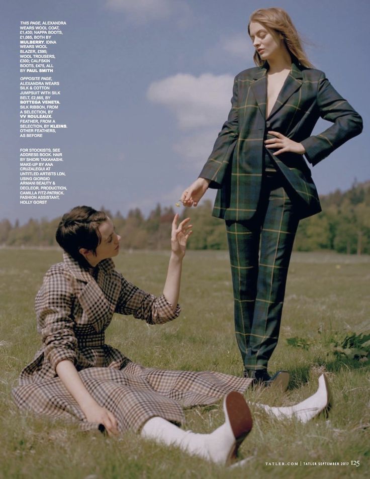 two women are standing in a field. one woman is wearing a plaid suit, the other is wearing a plaid coat. the woman in the suit is smiling, while the woman in the coat is looking at something.