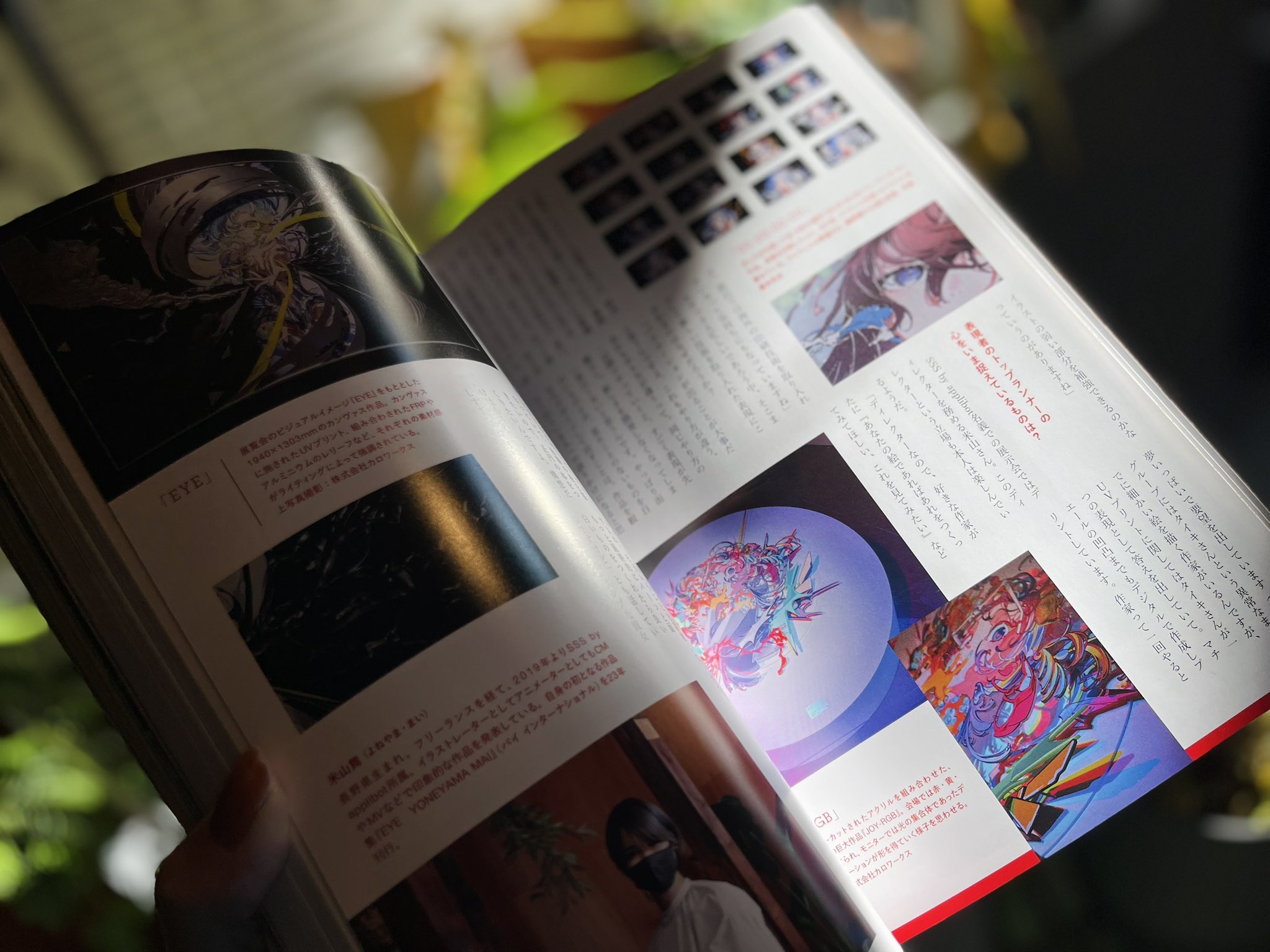 a firstperson perspective of someone holding open a japanese artbook or magazine. the pages showcase vibrant and detailed animestyle illustrations, with accompanying japanese text, likely an interview or artist feature. the lighting suggests a sunny indoor setting, with natural light casting shadows across the open book.