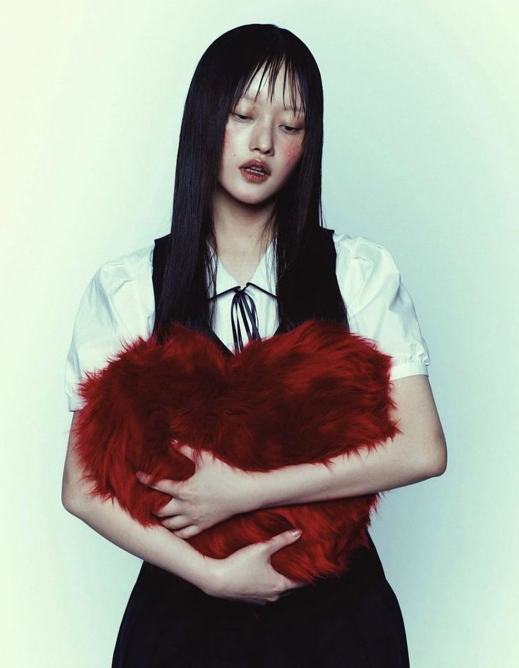 the image shows a young asian woman, possibly a model, posing in a studio setting against a lightcolored backdrop. she has long, straight black hair with bangs, pale skin, and subtle blush makeup.  she's wearing a white collared shirt over a black dress and vest. the most striking element is her pose: she's holding a large, heartshaped red fur pillow close to her chest, her expression suggesting sadness or melancholy.  the overall mood is quiet and introspective, with a soft focus enhancing the emotional tone. the image is likely a fashion photograph, emphasizing both the model's look and the visual symbolism of the heartshaped pillow.