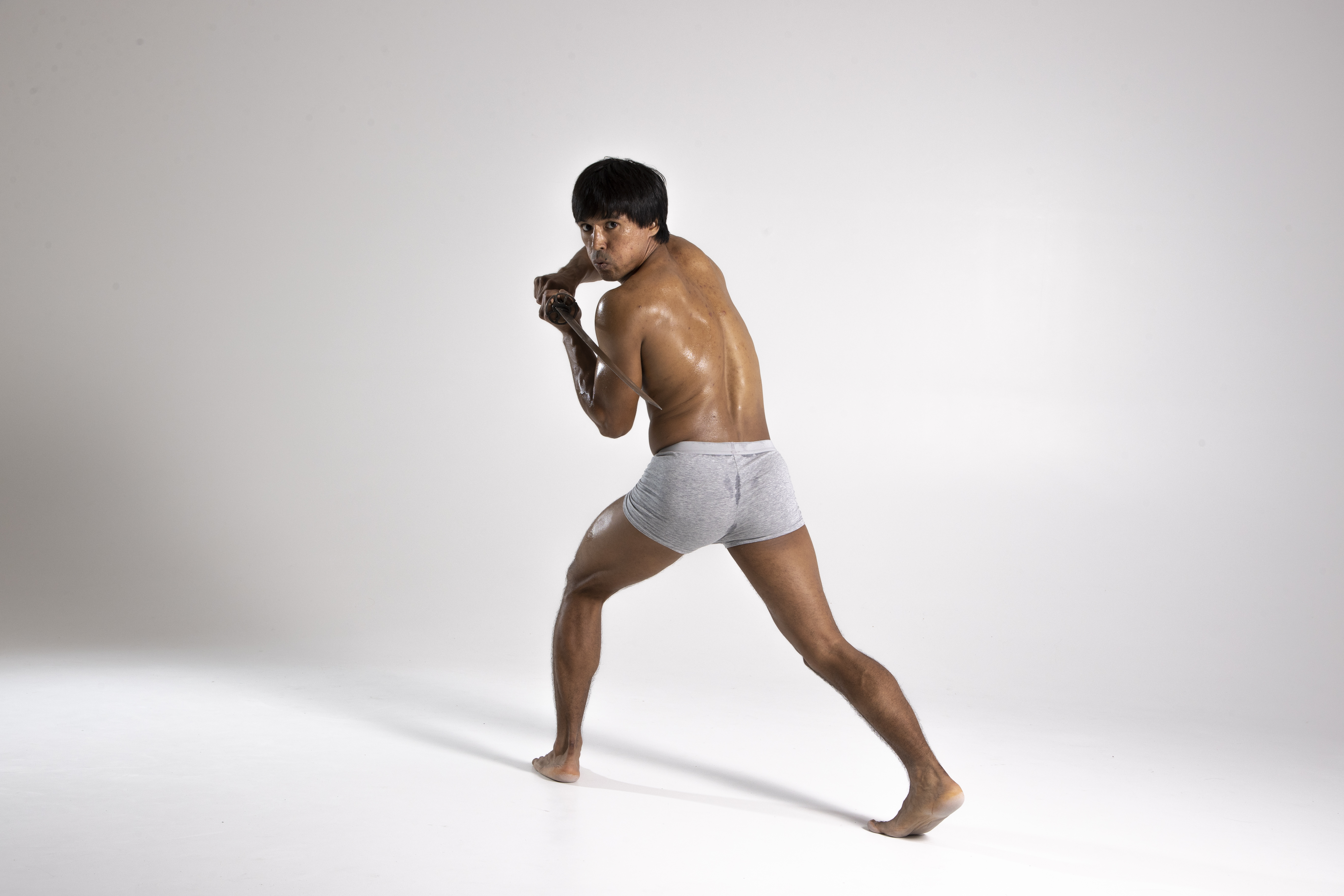 a shirtless man wearing gray shorts is holding a sword, looking back and posing in a fighting stance. he is barefoot on a white floor in front of a white background.